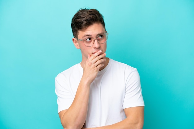 Joven apuesto brasileño aislado de fondo azul con gafas y con dudas