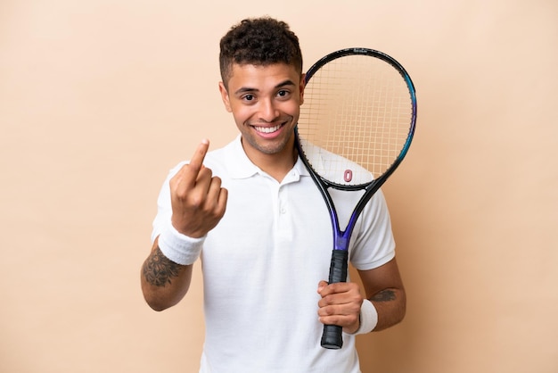 Joven apuesto brasileño jugando al tenis aislado de fondo beige haciendo el gesto de venir