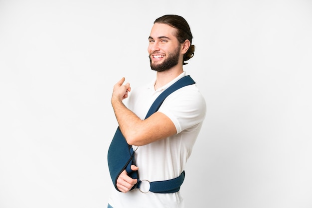 Un joven apuesto con un brazo roto y un cabestrillo sobre un fondo blanco aislado apuntando hacia atrás