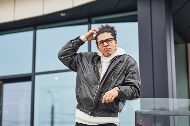 Un joven apuesto con cabello negro rizado y gafas está en la calle contra el edificio.