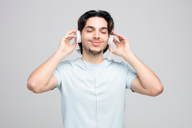 un joven apuesto complacido con auriculares agarrándolos escuchando música con los ojos cerrados aislado de fondo blanco