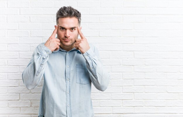 Joven apuesto contra una pared de ladrillos se centró en una tarea, manteniendo los dedos índice apuntando la cabeza.