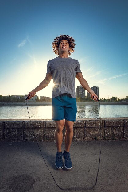 Foto joven y apuesto deportista saltando la cuerda cerca del río contra el cielo azul con la luz del amanecer.