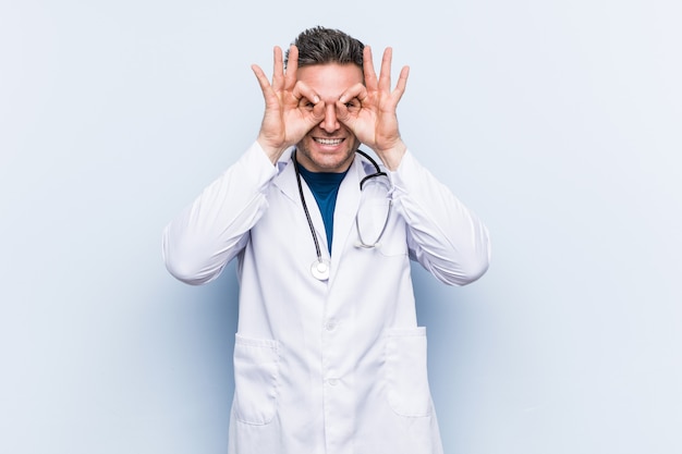 Joven apuesto doctor hombre mostrando signo bien sobre los ojos