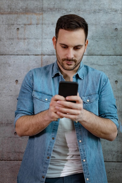 Joven apuesto empresario exitoso escribiendo un mensaje en un teléfono inteligente frente a un muro de hormigón.