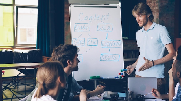 Un joven apuesto está haciendo una presentación creativa para el equipo usando una pizarra, está apuntando al texto con un marcador y hablando Entrenador de negocios y concepto de oficina