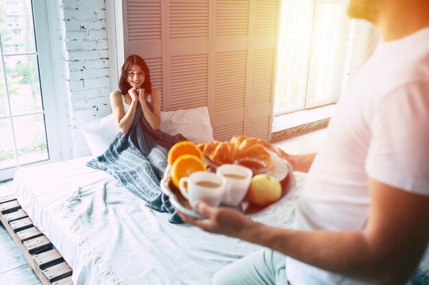 Un joven apuesto está parado con un desayuno romántico en la bandeja, lo hizo para su hermosa y linda chica o esposa sonriente, que está acostada en la cama y esperándolo