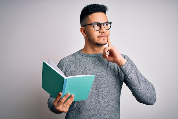 Joven apuesto estudiante inteligente leyendo un libro sobre fondo blanco aislado cara seria pensando en la pregunta idea muy confusa