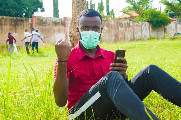 Joven apuesto hombre africano sentado afuera con mascarilla para prevenir, prevenir, prevenir el brote en la sociedad mientras opera su teléfono celular