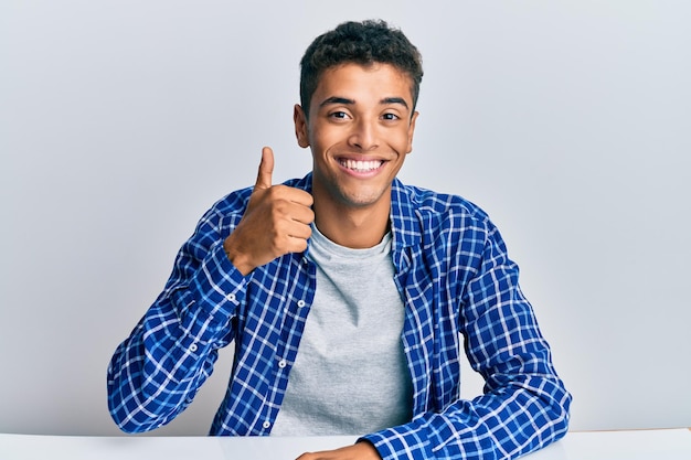 Joven apuesto hombre afroamericano con ropa informal sentado en la mesa sonriendo feliz y positivo con el pulgar hacia arriba haciendo excelente y signo de aprobación