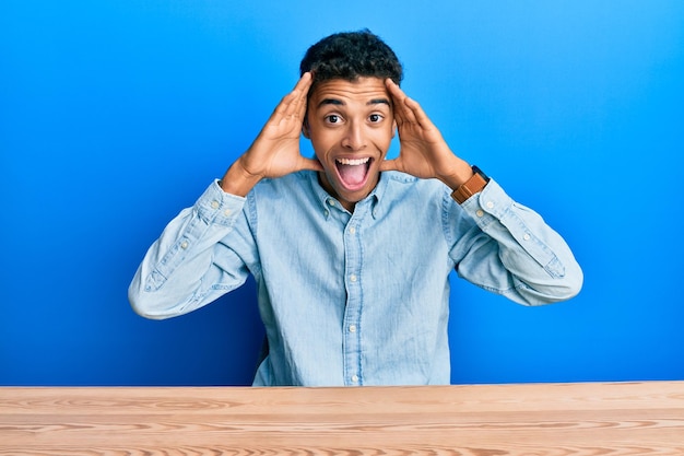 Joven apuesto hombre afroamericano vestido con ropa informal sentado en la mesa sonriendo alegre jugando a peek a boo con las manos mostrando la cara sorprendida y emocionada