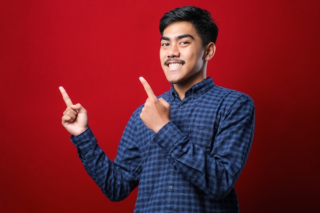 Joven apuesto hombre asiático con camisa casual sobre fondo rojo con una gran sonrisa en la cara, apuntando con el dedo de la mano hacia el lado mirando a la cámara.