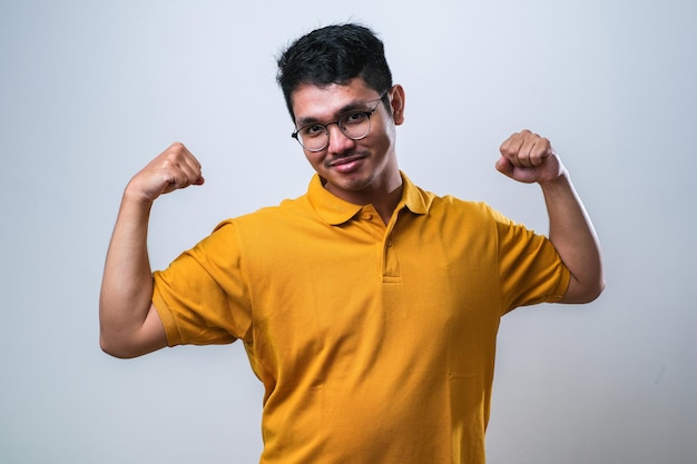 Joven apuesto hombre asiático sobre fondo blanco con ropa informal que muestra los músculos de los brazos sonriendo orgulloso concepto de fitness