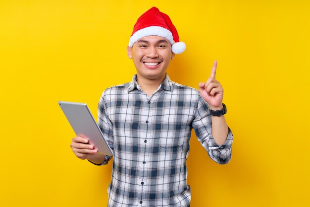 Joven apuesto hombre asiático sonriente con sombrero de Navidad sosteniendo una tableta apuntando con el dedo hacia arriba con una gran idea nueva sobre fondo amarillo Celebración Navidad y Feliz Año Nuevo 2023 concepto de vacaciones