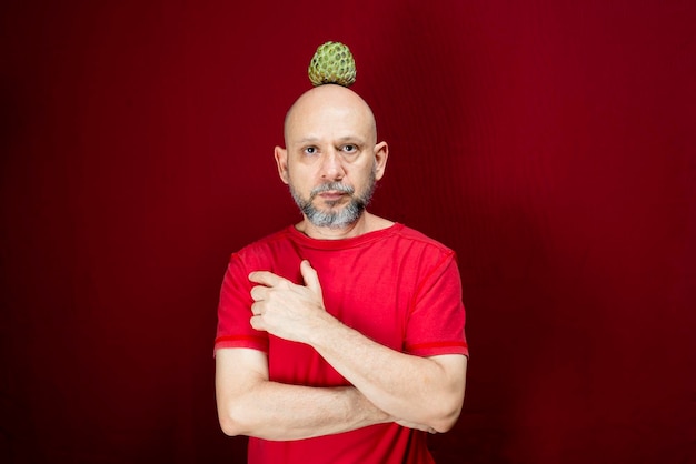 Foto joven apuesto hombre con barba, cabeza calva y camisa roja de pie con piña equilibrada en la cabeza contra fondo rojo persona positiva y sana