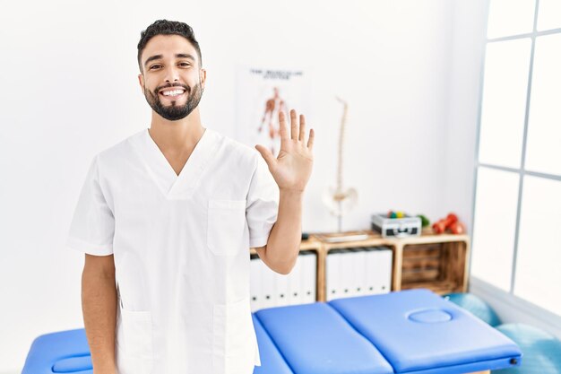 Joven apuesto hombre con barba trabajando en la clínica de recuperación del dolor que aparece y señala con los dedos número cinco mientras sonríe confiado y feliz.