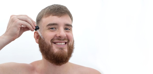 Joven apuesto hombre barbudo aplica loción de producto cosmético de aceite de suero para una hidratación saludable perfecta de la piel fresca crece la barba usando cuidado de la piel hidratante sobre fondo blanco