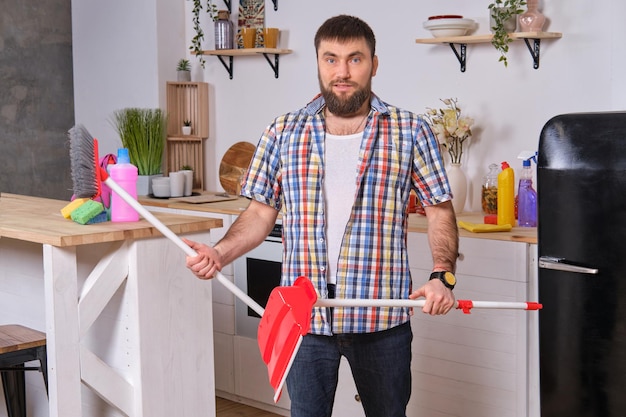 Joven apuesto hombre barbudo en la cocina con camisa a cuadros intenta lidiar con un recogedor y un ...