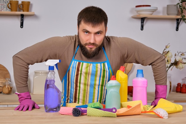 Joven apuesto hombre barbudo en la cocina, muestra a todo su personal de limpieza: detergentes, cepillos, aerosoles. Él cree que está listo para una limpieza real.