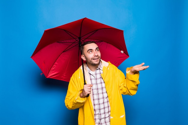 Joven apuesto hombre barbudo en impermeable amarillo con paraguas rojo aislado sobre fondo azul.