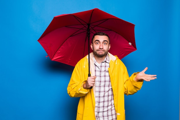 Joven apuesto hombre barbudo en impermeable amarillo con paraguas rojo aislado sobre fondo azul.