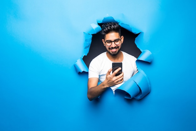 Joven apuesto hombre barbudo indio escribiendo mirada de teléfono desde el agujero de papel azul