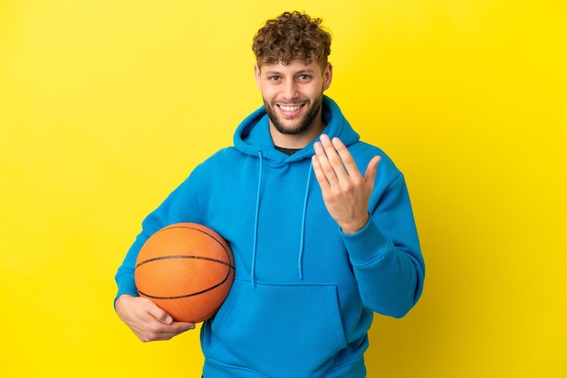 Joven apuesto hombre caucásico aislado de fondo amarillo jugando baloncesto y haciendo el gesto de venir