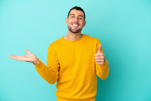 Joven apuesto hombre caucásico aislado de fondo azul con copyspace imaginario en la palma de la mano para insertar un anuncio y con los pulgares hacia arriba