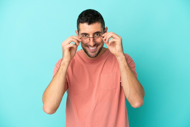 Joven apuesto hombre caucásico aislado de fondo azul con gafas con expresión feliz