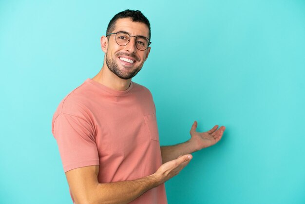 Joven apuesto hombre caucásico aislado de fondo azul con gafas y presentando algo