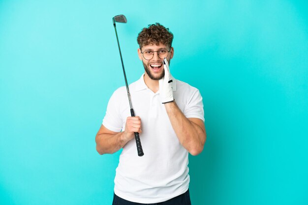 Joven apuesto hombre caucásico aislado de fondo azul jugando al golf y gritando con la boca abierta