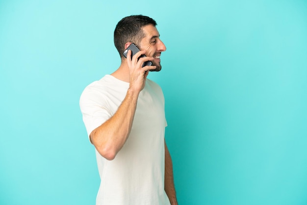 Foto joven apuesto hombre caucásico aislado de fondo azul manteniendo una conversación con el teléfono móvil con alguien