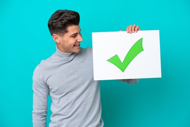 Joven apuesto hombre caucásico aislado en un fondo azul sosteniendo un cartel con texto Icono de marca de verificación verde con expresión feliz