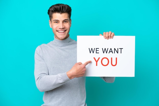 Joven apuesto hombre caucásico aislado en un fondo azul sosteniendo el tablero We Want You y apuntándolo