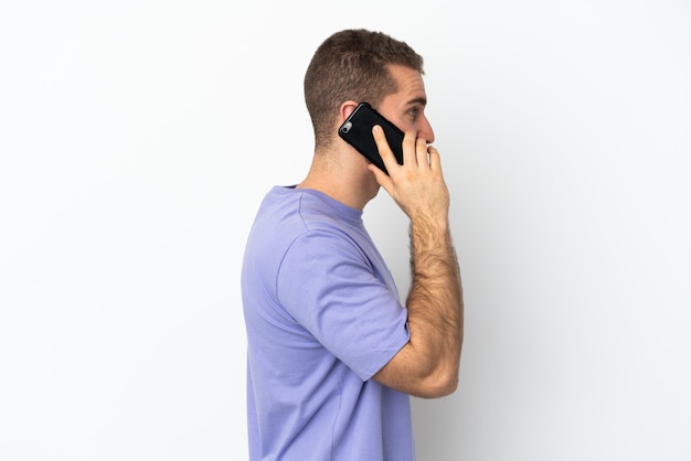 Joven apuesto hombre caucásico aislado en la pared blanca manteniendo una conversación con el teléfono móvil con alguien