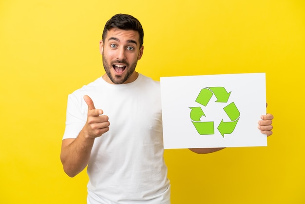 Joven apuesto hombre caucásico aislado sobre fondo amarillo sosteniendo un cartel con el icono de reciclaje y apuntando hacia el frente