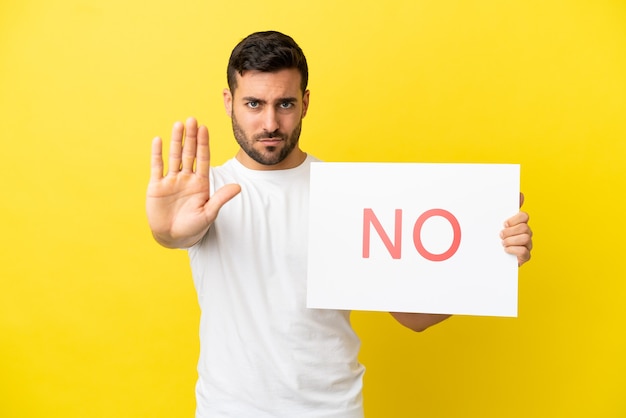 Foto joven apuesto hombre caucásico aislado sobre fondo amarillo sosteniendo un cartel con el texto no y haciendo señal de stop