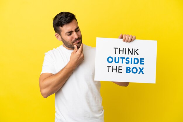 Joven apuesto hombre caucásico aislado sobre fondo amarillo sosteniendo un cartel con texto pensar fuera de la caja y pensar
