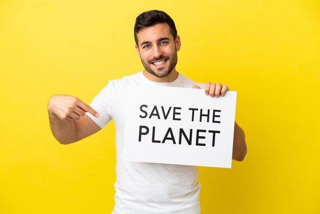 Joven apuesto hombre caucásico aislado sobre fondo amarillo sosteniendo un cartel con el texto Save the Planet y apuntando hacia él