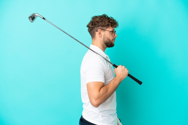 Joven apuesto hombre caucásico aislado sobre fondo azul jugando al golf en posición lateral