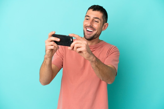 Joven apuesto hombre caucásico aislado sobre fondo azul jugando con el teléfono móvil