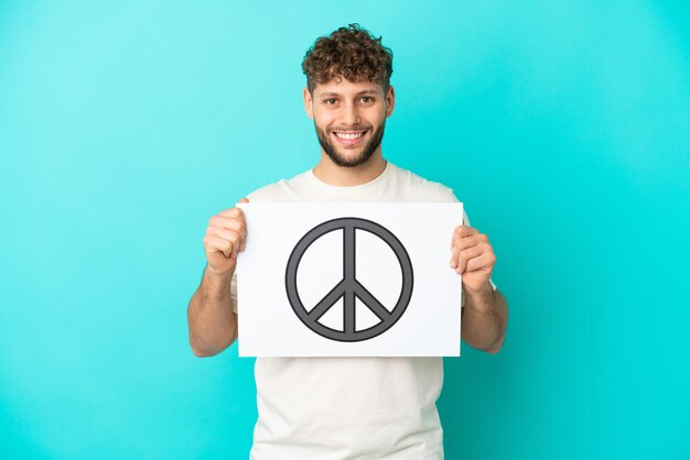 Joven apuesto hombre caucásico aislado sobre fondo azul sosteniendo un cartel con el símbolo de la paz con expresión feliz