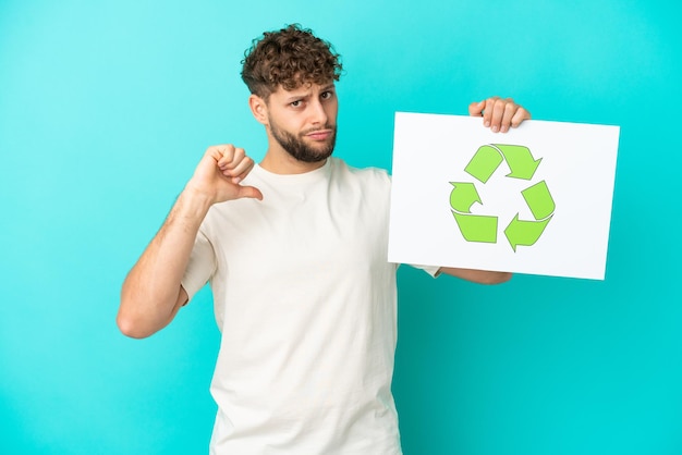 Joven apuesto hombre caucásico aislado sobre fondo azul sosteniendo una pancarta con icono de reciclaje con gesto orgulloso