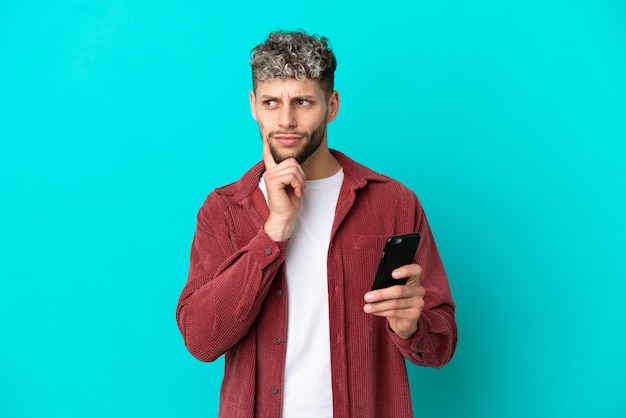 Joven apuesto hombre caucásico aislado sobre fondo azul con teléfono móvil y pensando