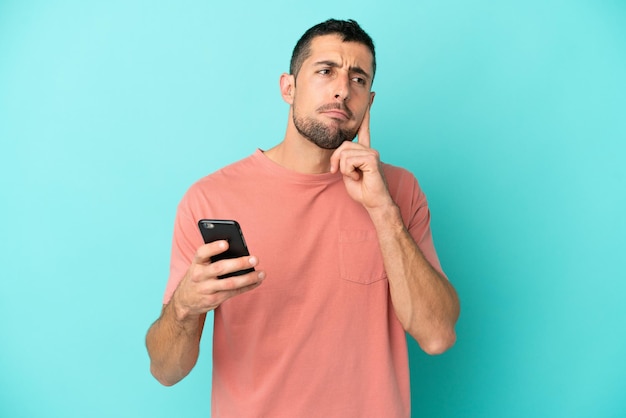 Foto joven apuesto hombre caucásico aislado sobre fondo azul con teléfono móvil y pensando