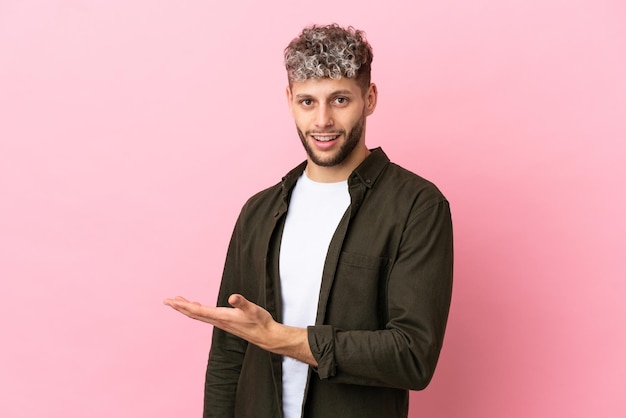 Joven apuesto hombre caucásico aislado sobre fondo rosa presentando una idea mientras mira sonriendo hacia