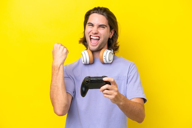 Joven apuesto hombre caucásico jugando con un controlador de videojuegos aislado en un fondo amarillo celebrando una victoria