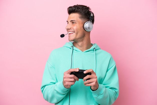 Joven apuesto hombre caucásico jugando con un controlador de videojuegos aislado en un fondo rosa mirando hacia un lado y sonriendo