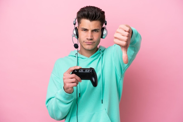 Joven apuesto hombre caucásico jugando con un controlador de videojuegos aislado en un fondo rosa mostrando el pulgar hacia abajo con expresión negativa