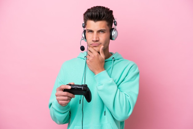 Joven apuesto hombre caucásico jugando con un controlador de videojuegos aislado en un pensamiento de fondo rosa